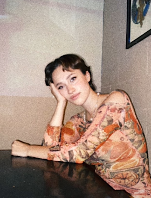 Person with light skin and short brown hair leaning on table with head in hand and orange-peach flowered shirt.