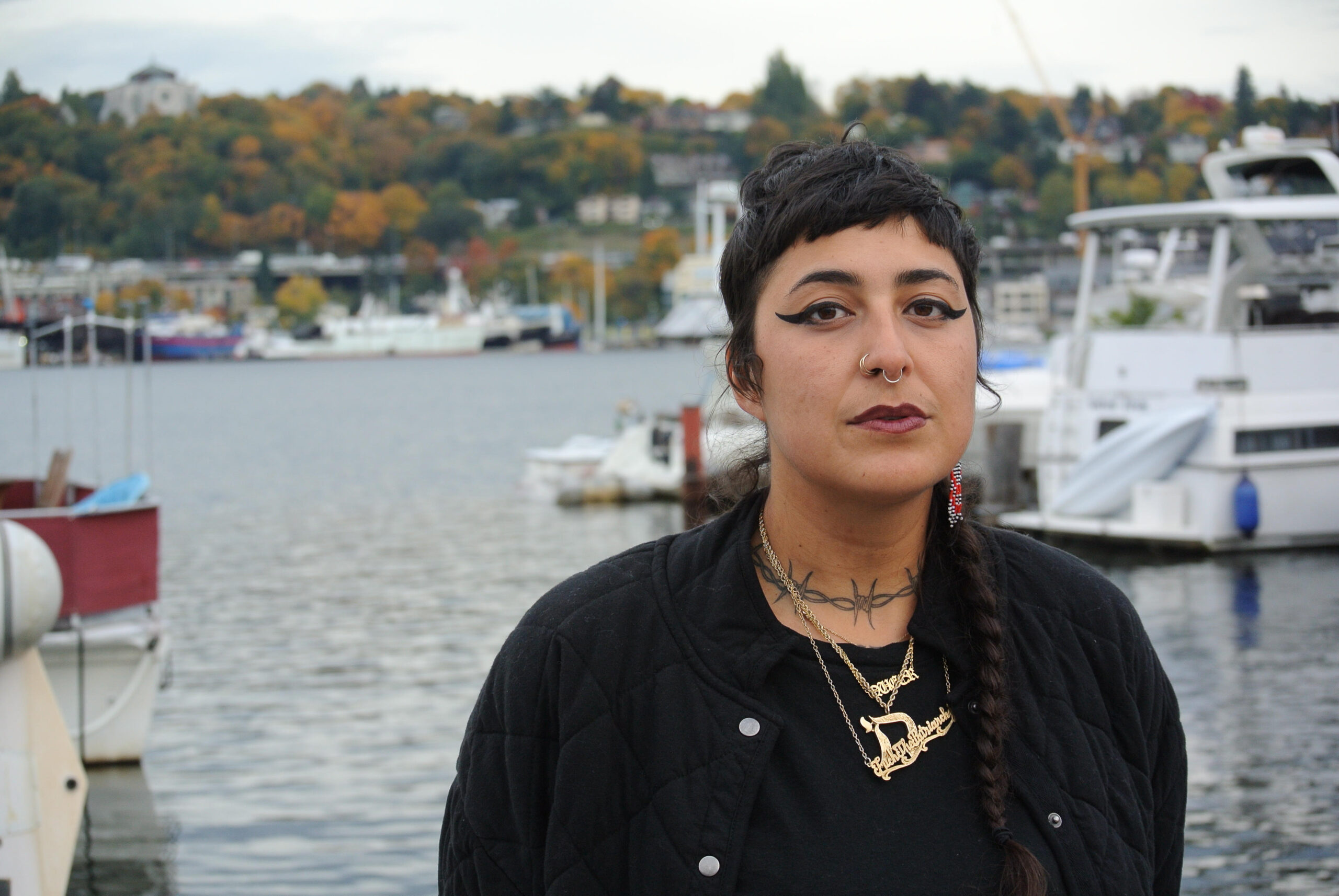 Person with dark hair and dark shirts and lots of necklaces standing in front of water and boats