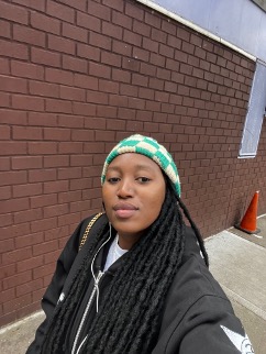 Selfie of dark skinned person with long black dreads wearing a white and green check knit cap and black coat in front of a red brick wall.