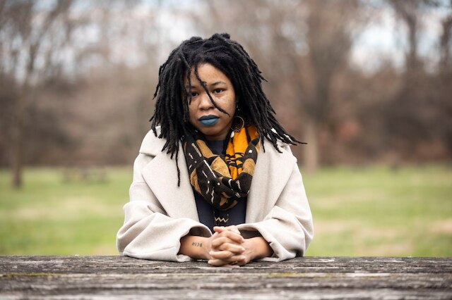 Person with dark hair in dreadlocks and white coat and colorful scarf with blue lipstick