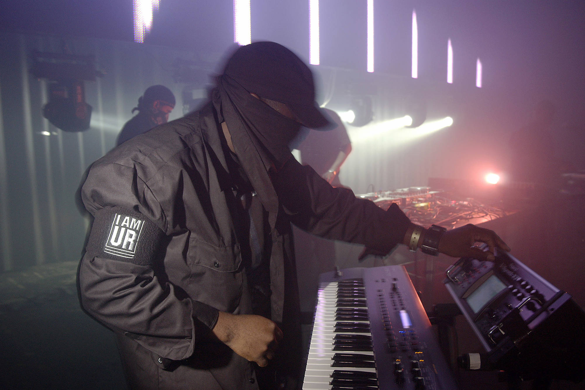 A person all in black with a black cap and black bandana mask is playing on synthesizer.