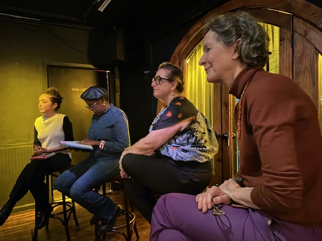 four people facing left on stools, the first in brown shirt and purple pants, than a black patterned top and balck pants with glasses, followed by a person in a cap, glasses and dark jean shirt and jeans and farthest away someone in a black and white top and black pants.