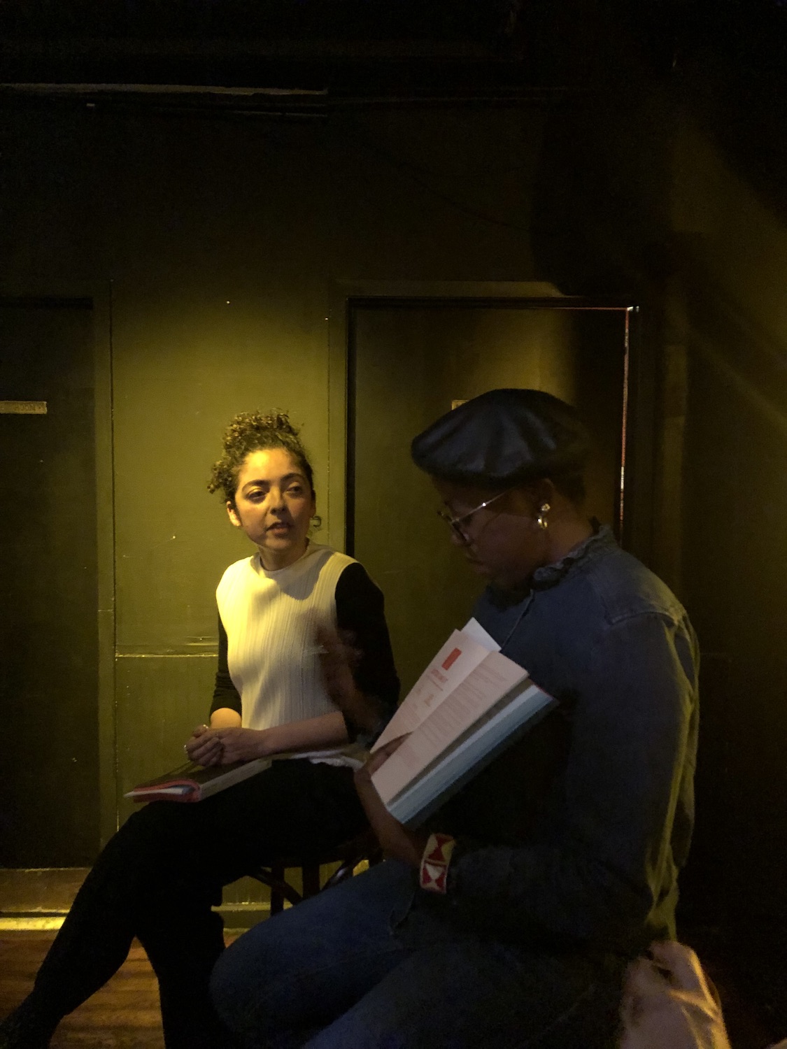 Two figures, the nearer video is holding up a book and wears a black cap, a dark denim shirt and glassees. The father person has a white shirt with black sleeves on and dark pants.