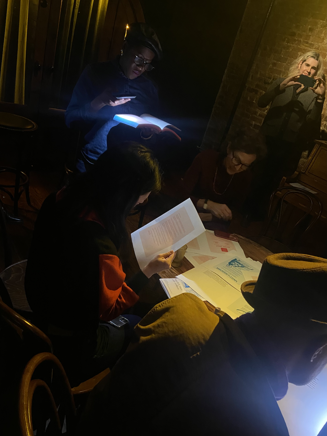 In the foreground are the backs of two people's heads who are looking at papers and farther away is a person in a dark cap with glasses holding a phone flashlight to read from the book in their hand.